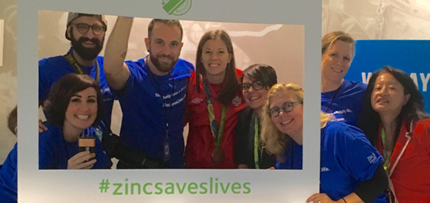Group of people in colorful shirts holding a frame with '#zincsaveslives' text at the bottom.